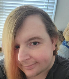 a white trans woman with medium length blonde-brown hair smiling in a selfie